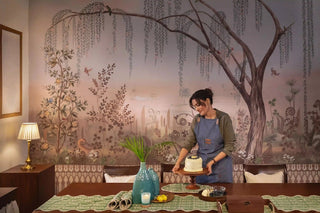 A woman in an apron setting a blueberry cake on a stand in front of a willow scenic mural