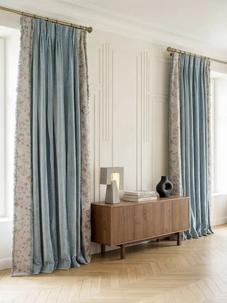 A complete lifestyle perspective featuring two panels of the Eternal Garden curtains framing a large window in a beautifully curated, modern-classic room. The drapes highlight a subtly textured, light blue center flanked by vintage floral borders and delicate fringed edges. They hang elegantly from brass-accented rods above a warm wooden credenza.