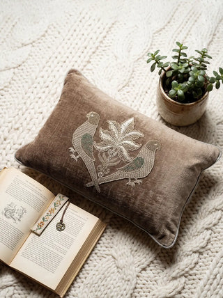 A flatlay view of the Jewelled Parrot velvet cushion with parakeet embroidery, styled with a book and plant.