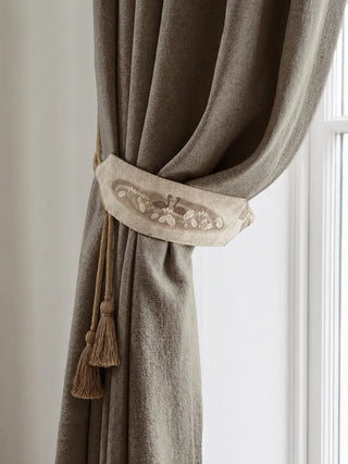 Kumud is shown elegantly holding a textured taupe curtain in place beside a bright window. The wide linen tieback features a soft vintage motif of two swans among water lilies and lotus flowers, paired with thick twisted cords and hanging tassels. The overall look feels warm, rustic, and graceful, adding a handcrafted storybook charm to the curtain setting.