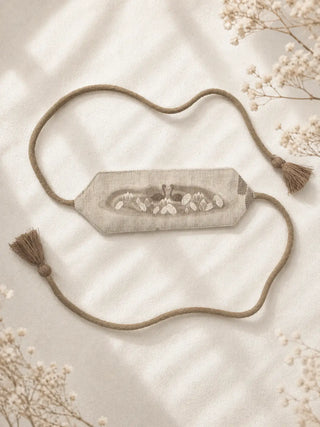 Kumud is displayed as a standalone curtain tieback on a soft ivory fabric background. Its structured linen band highlights the central swan, lotus, and water lily design, while the twisted rope extensions and matching tassels frame the piece beautifully. The muted oatmeal and taupe tones create a calm, vintage-inspired look with natural textile elegance.