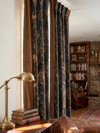 A full-length lifestyle perspective of the Majestica curtains, showcasing their rich drape in a curated library study. The deep blue fantasy print center is framed by wide brown velvet borders, positioned next to a dark wood bookshelf, a leather armchair, a stone fireplace, and a vintage brass lamp.