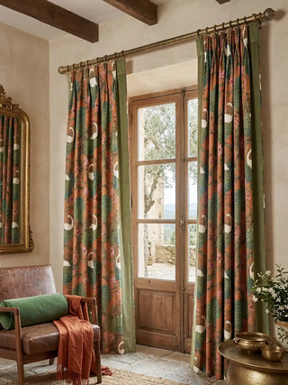 A full lifestyle perspective showcasing two panels of the Silk Route curtains framing rustic wooden doors in a sun-drenched, Mediterranean-inspired room. The rich ochre botanical print and plush sage green velvet borders perfectly complement the warm, earthy tones of the leather armchair, brass curtain rod, and antique gold accents.