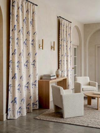 A complete lifestyle perspective featuring two panels of the Stallion curtains in a bright, contemporary living space or entryway. The drapes feature a natural beige linen background adorned with ink-blue horse illustrations and polka dots. Distinctive ink-blue side loops run vertically down the outer edges, complementing the minimalist, neutral decor and warm wood console table.