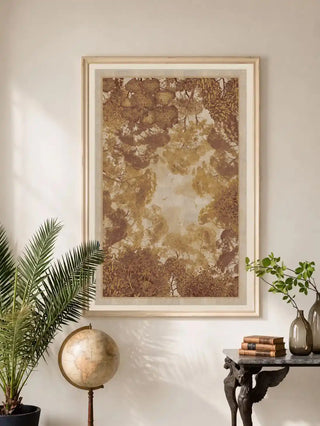The framed artwork is mounted on a neutral beige wall above a globe, potted palm plant, and a side table with stacked books. Soft sunlight casts gentle shadows, creating an elegant, relaxed study environment.