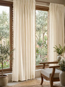 full lifestyle perspective featuring two panels framing a large wooden window that looks out onto a lush garden. The airy off-white linen softly filters the natural light, while the delicate tassel lace trim along the...