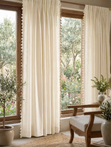 full lifestyle perspective featuring two panels framing a large wooden window that looks out onto a lush garden. The airy off-white linen softly filters the natural light, while the delicate tassel lace trim along the...