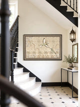 The Birds of Anqa embroidered wall art framed on a wall beneath a black-railed staircase beside a console table and lantern sconce.