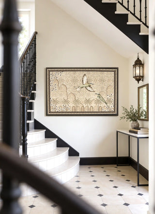 The Birds of Anqa embroidered wall art framed on a wall beneath a black-railed staircase beside a console table and lantern sconce.