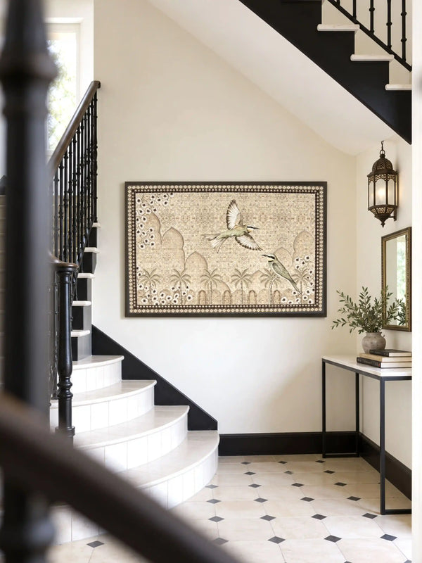 The Birds of Anqa embroidered wall art framed on a wall beneath a black-railed staircase beside a console table and lantern sconce.