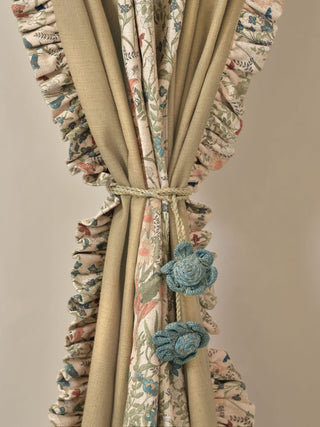 A detailed close-up of the curtain elegantly tied back in the center. The image highlights the beautiful layering of the solid linen side panel alongside the intricate floral print, beautifully framed by a matching printed floral ruffle. A charming handcrafted floral tie-back in a contrasting soft blue adds a touch of whimsical detail.