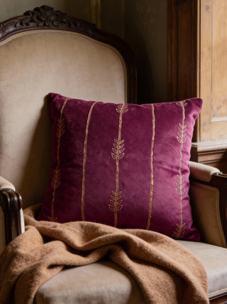 A luxury plum velvet cushion with gold-embroidered vine stripes styled on a classical beige armchair with a tan throw.