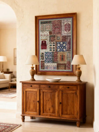 A framed Aalishan art piece displayed on a cream wall above a rustic wooden sideboard with ivory lamps.