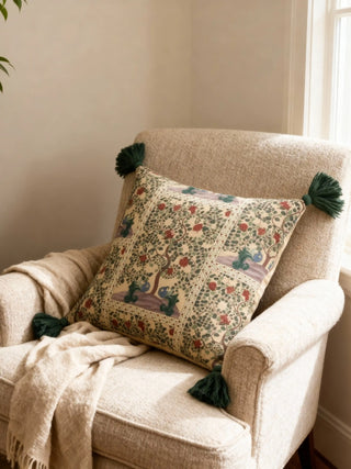 A luxury Akriti cushion cover featuring a Pichwai-style garden scene and deep green corner tassels on a neutral textured armchair.