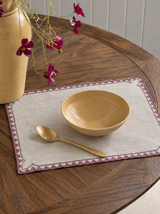 An Amrah table mat from the set in Natural & Mauve on a round wooden table, set with a stoneware bowl, a brass spoon and a trailing floral arrangement.