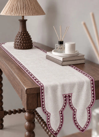 The Amrah Runner in Natural & Mauve dresses a wooden side table beside a rattan lampshade, a reed diffuser and stacked books.