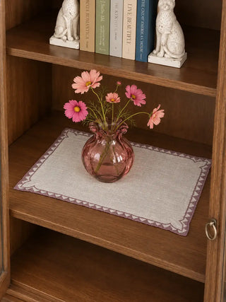 Amrah Table Mats in Natural & Mauve laid on a wooden shelf styled with a pink glass vase of cosmos flowers and stacked books.