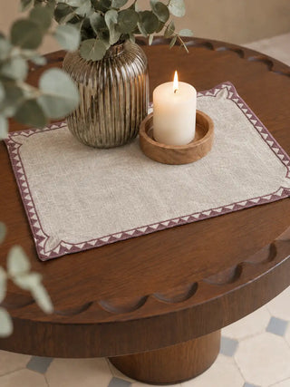 Amrah Table Mats in Natural & Mauve on a round carved wood side table, styled with a lit pillar candle and a ribbed glass vase of eucalyptus.