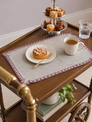 Amrah Table Mats in Natural & Mauve set on a wooden tea cart with a tiered cake stand, a plated dessert and a mug of tea.