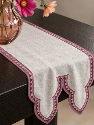 The Amrah Table Runner in Natural & Mauve drapes over a dark wood console beside a pink glass vase, showing its cusped double-point end.