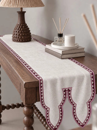 The Amrah Table Runner in Natural & Mauve dresses a wooden side table beside a rattan lampshade, a reed diffuser and stacked books.