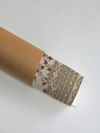 The Anant Vriksha wall art print partly unrolled from its shipping tube, showing a fruiting vine border beside a diamond-lattice field in red, black and tan.