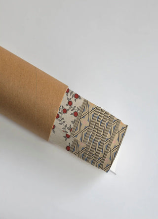The Anant Vriksha wall art print partially unrolled from its kraft paper shipping tube, showing a fruiting vine border beside a geometric diamond-lattice field in red, black and tan.