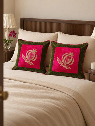 A pair of Anarkali cushion covers on a cream bed with a dark wood headboard, gold-embroidered pomegranates on fuchsia velvet framed in olive.