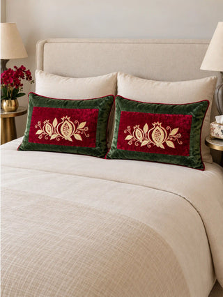 A pair of Annar Bagh lumbar cushions on a cream bed with linen pillows, gold-embroidered pomegranates on red velvet inside olive green borders.