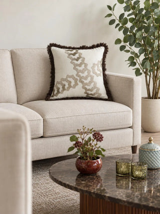 Aurea cushion cover with champagne embroidery and brown fringe on a cream textured sofa, beside a tall leafy plant in a speckled pot and a marble-topped coffee table with a glazed bowl of berry stems.