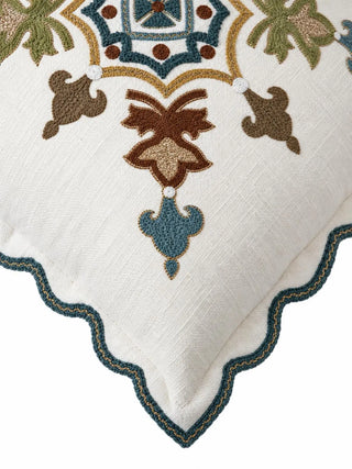 A close-up of the Avsar cushion cover corner showing dense looped stitching in teal, rust and olive with gold-toned outlines, and a teal braid-edged scalloped flange on a slubbed ivory weave.