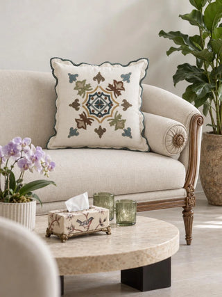 Avsar cushion cover with a multicolour embroidered medallion and a teal scalloped edge on a curved cream sofa with carved wood arms, beside a round stone-topped coffee table with a lilac orchid, green glass candle holders and a bird-print tissue box.