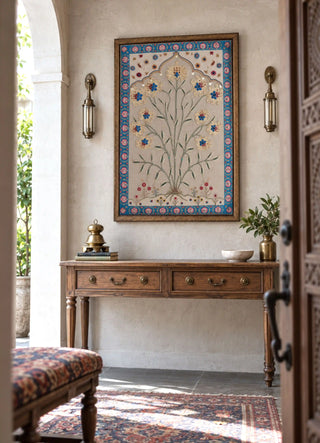 The Bagh-e-Noor embroidered wall art framed above a wooden console table between brass wall sconces beside an open entrance door.
