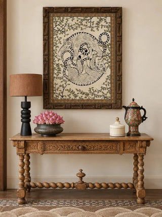 The Bagha Leela wall art in a carved wooden frame above a carved wooden console with a black lamp and pink flowers in a metal bowl.