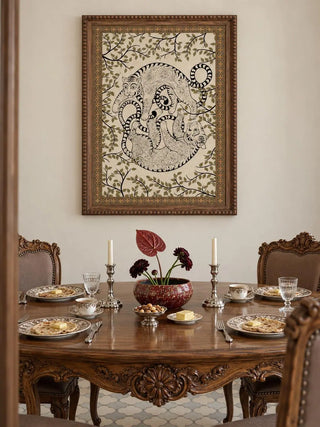 The Bagha Leela wall art in a beaded wooden frame above a round walnut dining table set with plates, candlesticks and a red bowl.