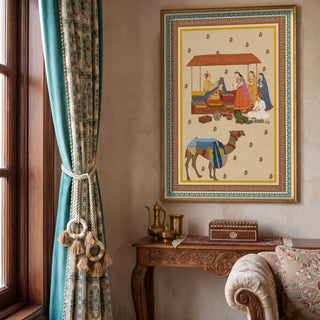 Bazar Heritage wall art in a gilt frame on a warm taupe limewash wall above a carved wooden console with brass dallah and decorative box, alongside teal block-print curtains with tassel tieback and a cream embroidered sofa