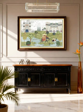 Bharat Mughal miniature wall art in sage ivory, framed in dark walnut with a gold fillet, hung above a carved black sideboard with silver elephant figurines and a crystal chandelier in a marble-floored living room.