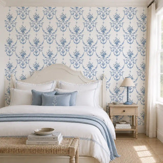 The Blue Garland wallpaper in Powder Blue & White installed on a feature wall in a bedroom, showing a rose bouquet and ribbon trellis pattern behind a cream headboard and wooden nightstand. view-bedroom