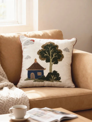 A luxury Celestial Village cushion cover with hand-embroidered village motifs and red piping resting on a tan velvet sofa.