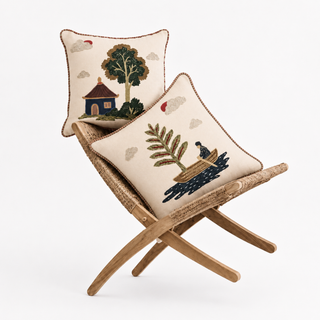 he Celestial Village Pine & Pagoda cushion set styled on a woven rattan bench, showing a chinoiserie-style cushion with an embroidered pagoda-roofed house and tree, and a second cushion with a pine branch and a figure in a boat, both on ivory cotton-linen with stylised clouds.