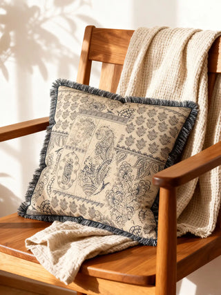A luxury Chhaya cushion cover featuring vintage charcoal paisley motifs and a fringed border resting on a wooden chair with a cream throw.