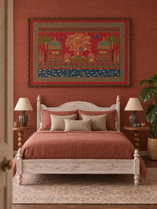 The Chitrakar wall art in a wavy dark wood frame on a terracotta wall above a whitewashed wooden bed with rust bedding and ribbed lamps.