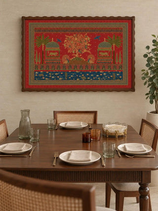 The Chitrakar wall art in a scalloped dark wood frame on a cream wall above a walnut dining table with cane-back chairs and ribbed glassware.
