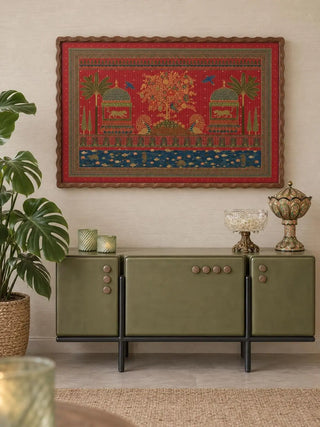 The Chitrakar wall art in a scalloped dark wood frame on a cream wall above an olive green sideboard with green glass votives, a lidded glass urn and a monstera plant.