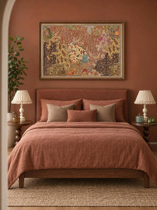 The Chitravali wall art in a carved gold frame on a terracotta wall above an upholstered bed with rust linen bedding and pleated white lamps.