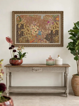 The Chitravali wall art in a carved gold frame above a whitewashed console with a red planter of orchids and anthuriums, beside a fiddle-leaf fig.