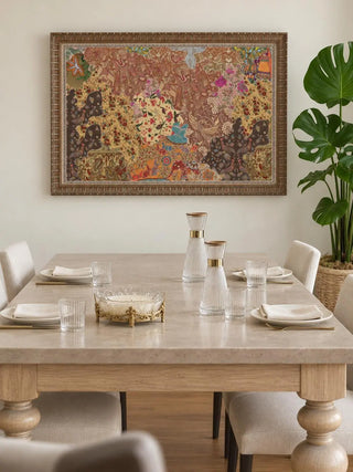 The Chitravali wall art in a carved gold frame above a stone-topped dining table with linen chairs, ribbed glass carafes and a monstera plant.