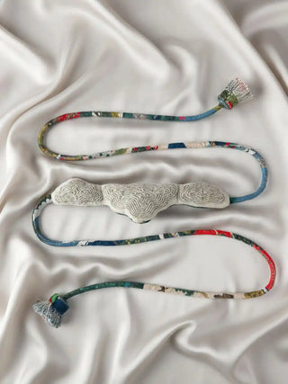 A flat lay view of the Cloud Loom curtain tie back on silk fabric, showcasing the full length of the variegated woven rope.