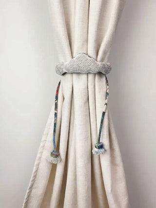 The Cloud Loom hand-embroidered cloud tie back gathering an ivory linen curtain panel against a clean white wall backdrop.