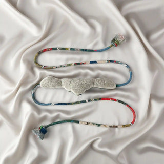 Flat lay of the Cloudloom Tassel showing the cloud-shaped embroidered central piece and the brightly colored braided cords on silk.